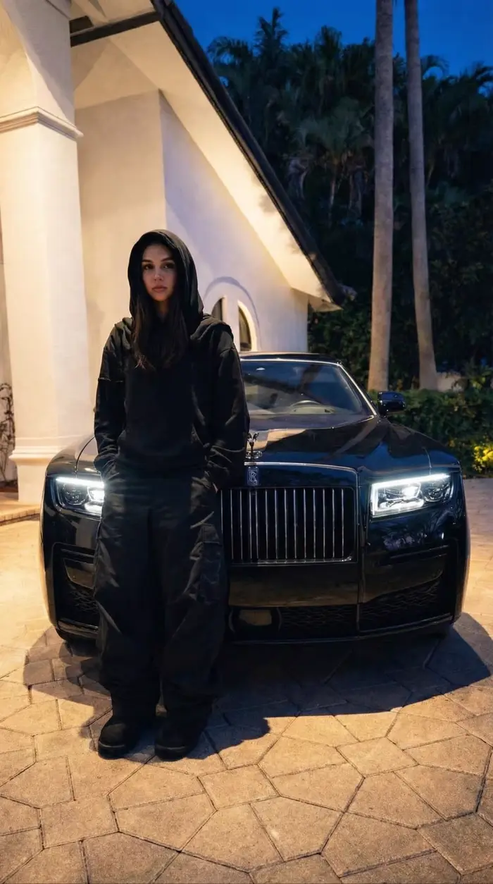A woman standing in front of a black Rolls-Royce at night.